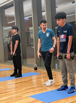Trainer observes two young men doing dumbbell exercises in a gym.